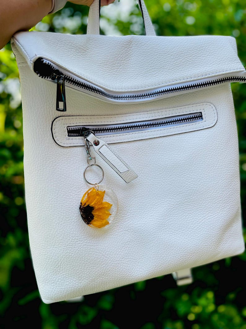 May include: White leather backpack with multiple zipped compartments. A clear resin keyring with a yellow sunflower is attached to the front zip. The backpack has a top flap and a handle.