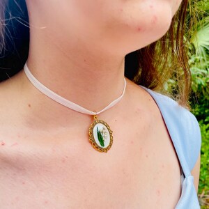 Pode incluir: Um colar choker de cetim branco com um pingente oval dourado com um design de flores prensadas.