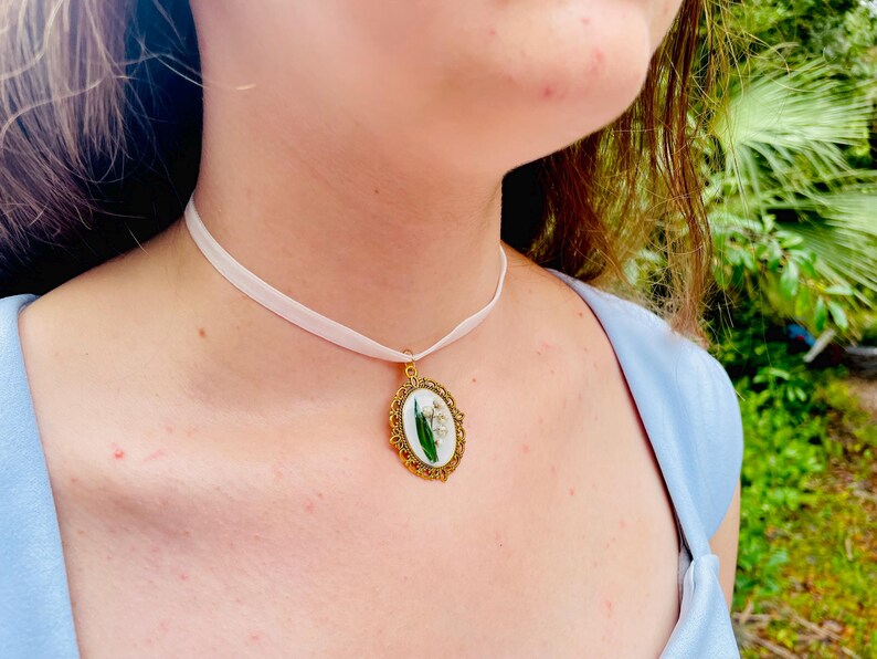 Pode incluir: Um colar choker de cetim branco com um pingente oval dourado com um design de flores prensadas.
