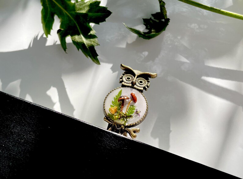 May include: A bronze owl brooch with a white background and a resin inlay of mushrooms and greenery.