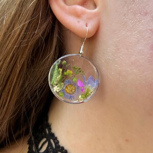 Puede incluir: Pendientes redondos transparentes con flores secas encapsuladas en resina. Los pendientes presentan una variedad de flores coloridas, incluyendo flores moradas, amarillas y rosas, sobre un fondo transparente. Cuelgan de ganchos plateados.