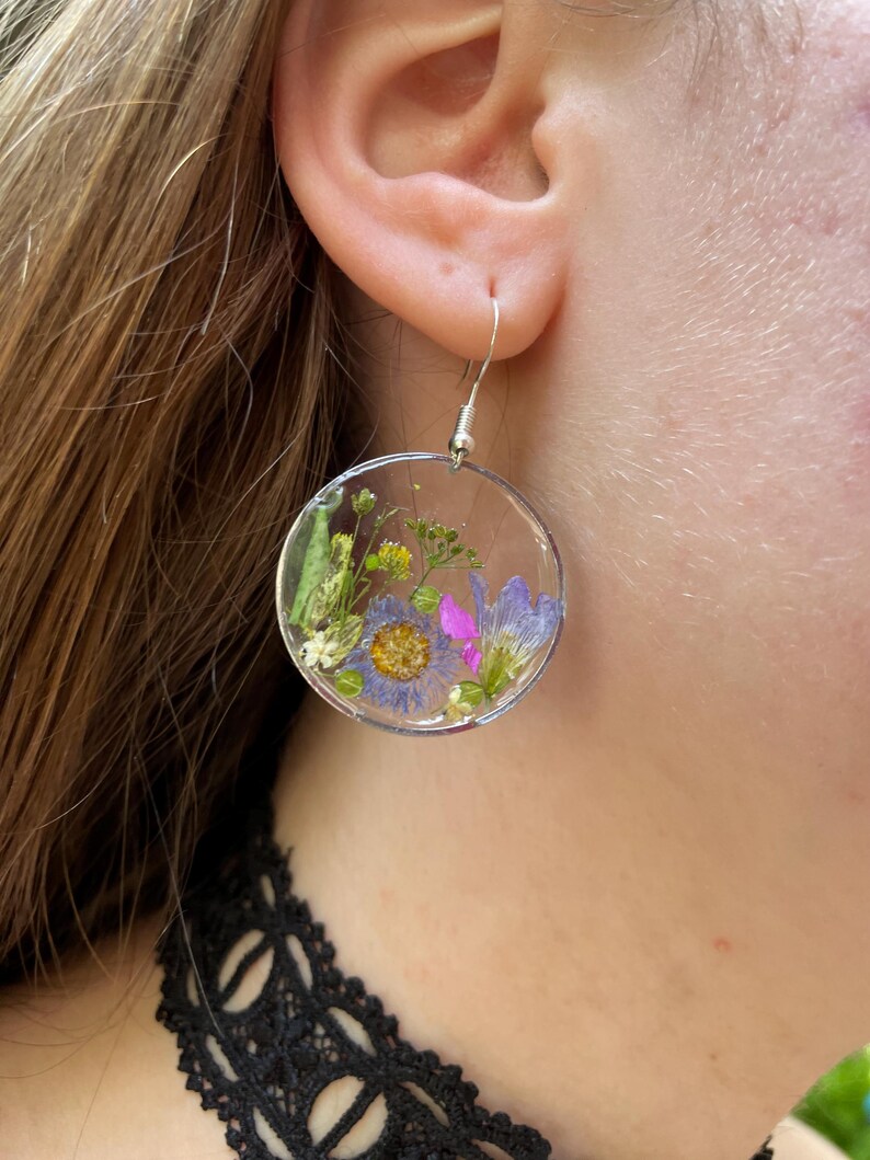 Puede incluir: Pendientes redondos transparentes con flores secas encapsuladas en resina. Los pendientes presentan una variedad de flores coloridas, incluyendo flores moradas, amarillas y rosas, sobre un fondo transparente. Cuelgan de ganchos plateados.
