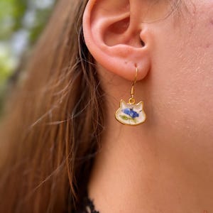 Puede incluir: Pendientes dorados con forma de gato con fondo blanco y dise&ntilde;o floral azul. Los pendientes cuelgan de un gancho dorado. Un accesorio &uacute;nico y elegante.