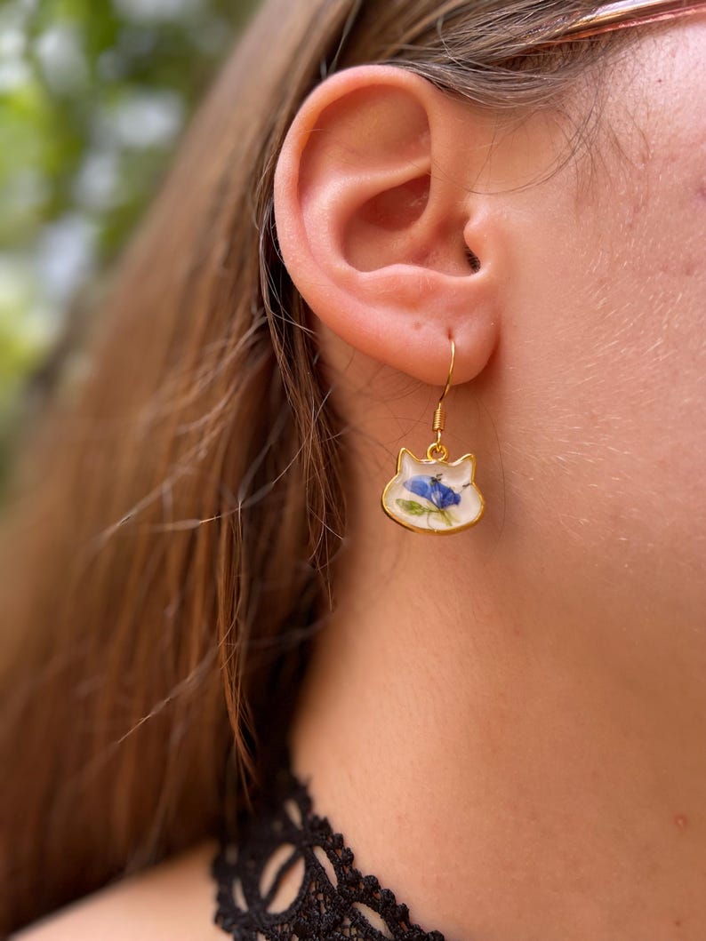 Puede incluir: Pendientes dorados con forma de gato con fondo blanco y dise&ntilde;o floral azul. Los pendientes cuelgan de un gancho dorado. Un accesorio &uacute;nico y elegante.