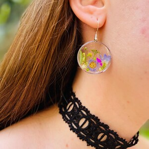 Puede incluir: Pendientes circulares transparentes con flores secas en tonos morados, amarillos y verdes. Los pendientes cuelgan de ganchos plateados. Se ve un collar de gargantilla de encaje negro.