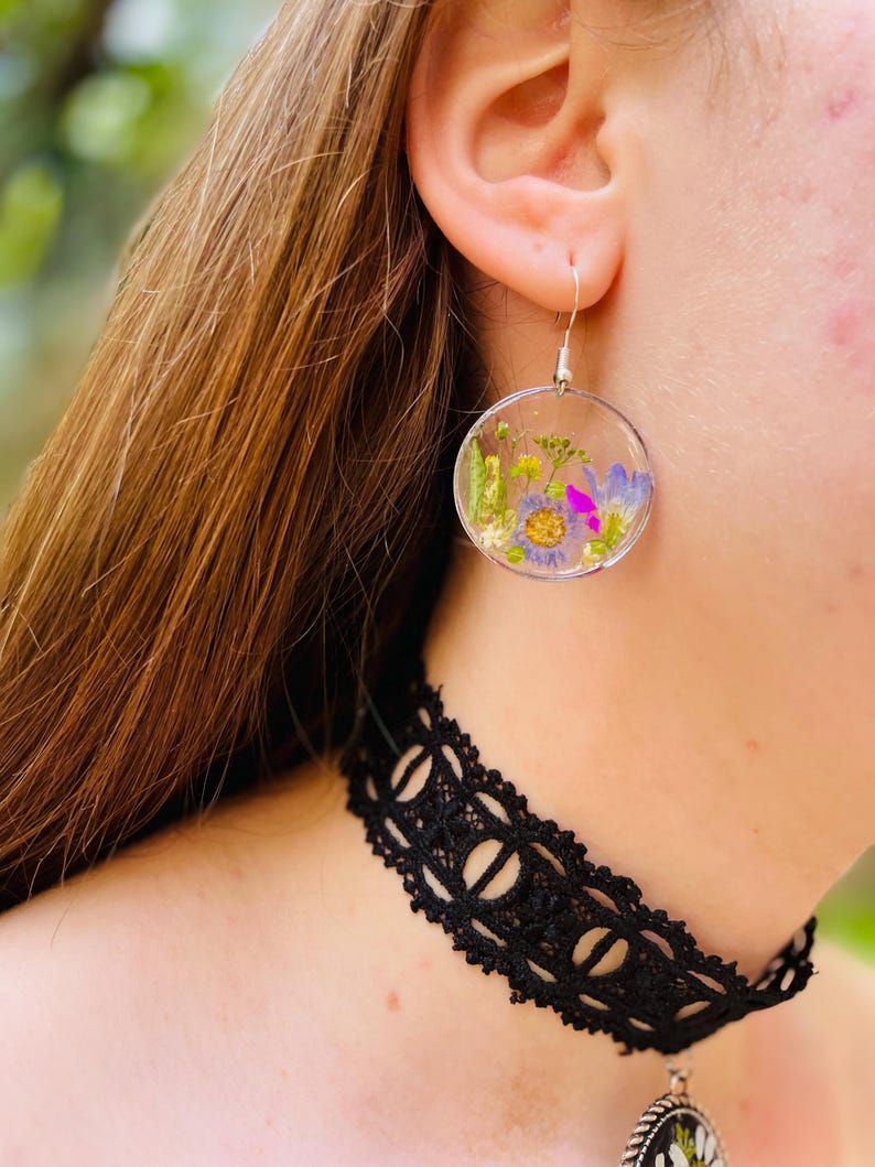 Puede incluir: Pendientes circulares transparentes con flores secas en tonos morados, amarillos y verdes. Los pendientes cuelgan de ganchos plateados. Se ve un collar de gargantilla de encaje negro.