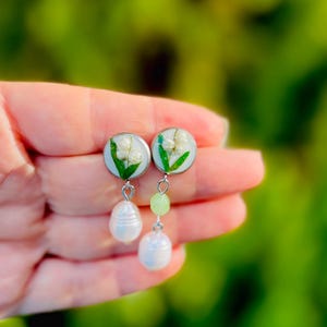 May include: A pair of handmade earrings featuring small, round, light blue resin settings with white flowers and green leaves. Each earring has a light green bead and a white pearl drop. The earrings are held in a hand.