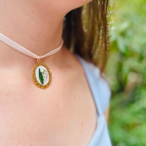 Pode incluir: Um colar de choker de cetim branco com um pingente oval dourado que apresenta uma flor de l&iacute;rio do vale prensada.