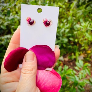 May include: A pair of heart-shaped earrings with pink and gold pressed flower accents. The earrings are displayed on a white card with a white background.