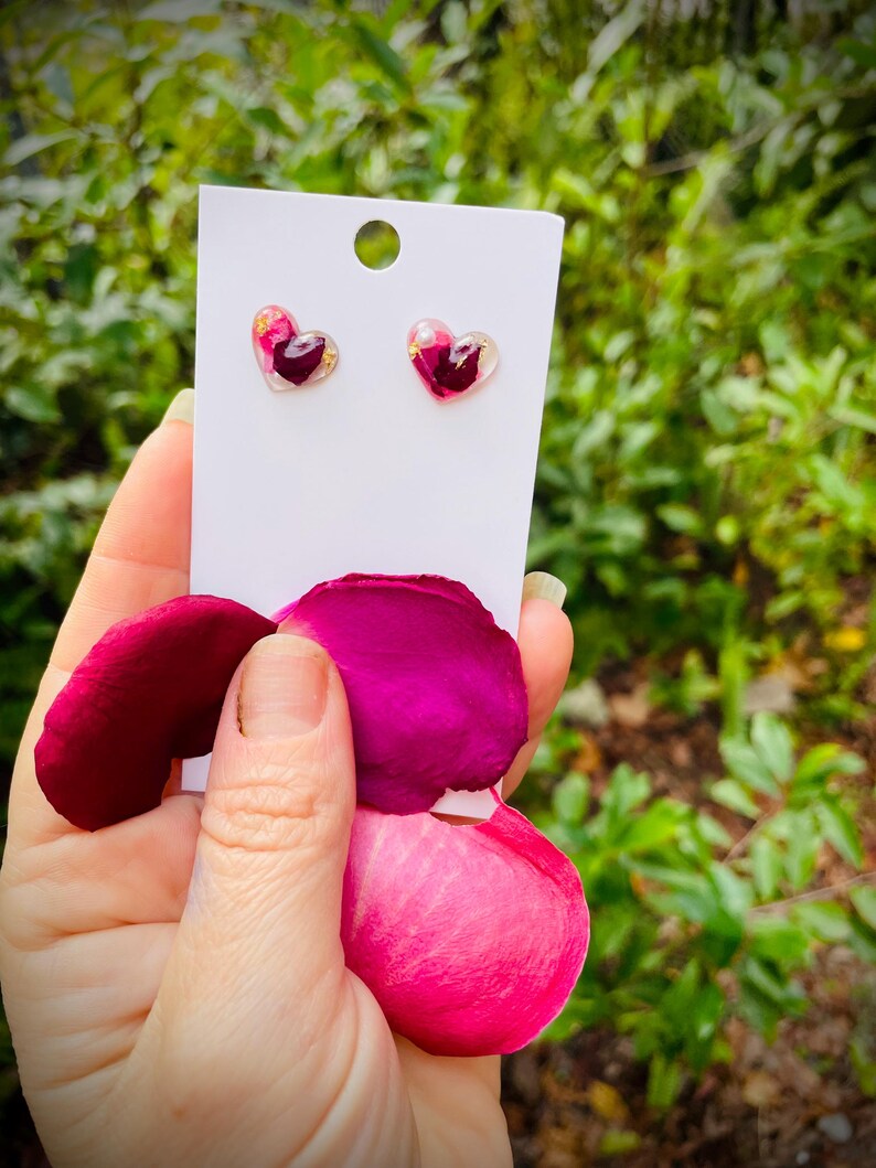 May include: A pair of heart-shaped earrings with pink and gold pressed flower accents. The earrings are displayed on a white card with a white background.