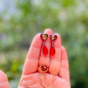 May include: A pair of red heart-shaped earrings with white floral details and dangling red teardrop charms. A red ring with a floral design is also visible in the image.