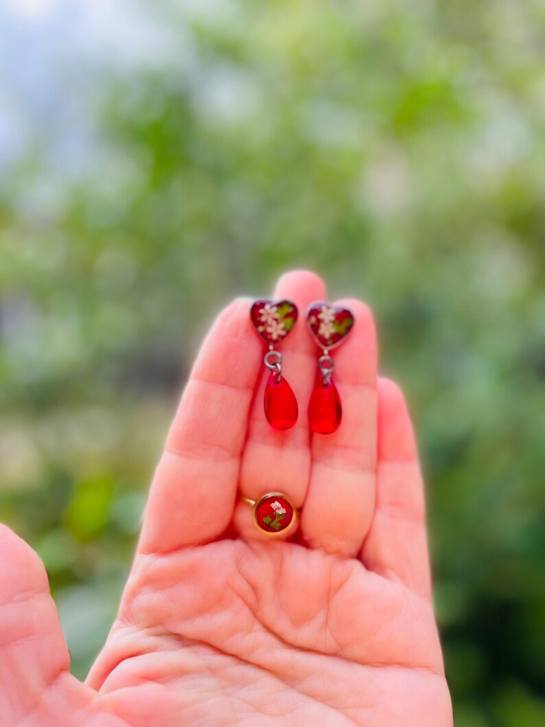 May include: A pair of red heart-shaped earrings with white floral details and dangling red teardrop charms. A red ring with a floral design is also visible in the image.
