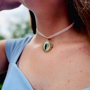 Pode incluir: Um colar de fita branca com um pingente oval dourado com uma flor de l&iacute;rio do vale prensada.