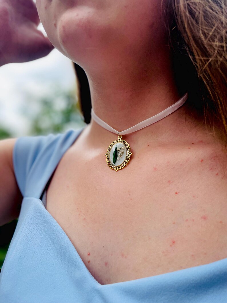 Pode incluir: Um colar de fita branca com um pingente oval dourado com uma flor de l&iacute;rio do vale prensada.