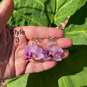 Pode incluir: Um par de brincos com flores de orqu&iacute;dea roxo-claras com centros roxo-escuros, cada um preso a um pequeno pingente de la&ccedil;o de metal dourado. O texto 'Style 3' &eacute; vis&iacute;vel no lado esquerdo da imagem, perto dos brincos.