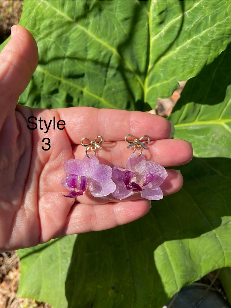 Pode incluir: Um par de brincos com flores de orqu&iacute;dea roxo-claras com centros roxo-escuros, cada um preso a um pequeno pingente de la&ccedil;o de metal dourado. O texto 'Style 3' &eacute; vis&iacute;vel no lado esquerdo da imagem, perto dos brincos.