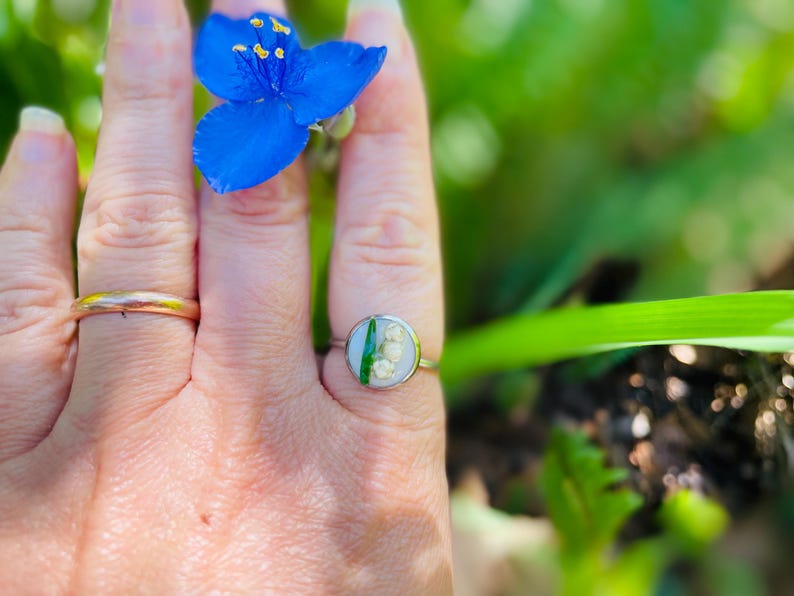 May include: A silver ring with a round, clear resin setting featuring a pressed lily of the valley flower and a green leaf.
