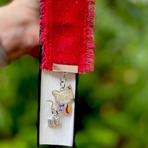 May include: A red velvet bookmark with a frayed edge and a silver charm of a cat holding a book. The cat is decorated with pressed flowers.