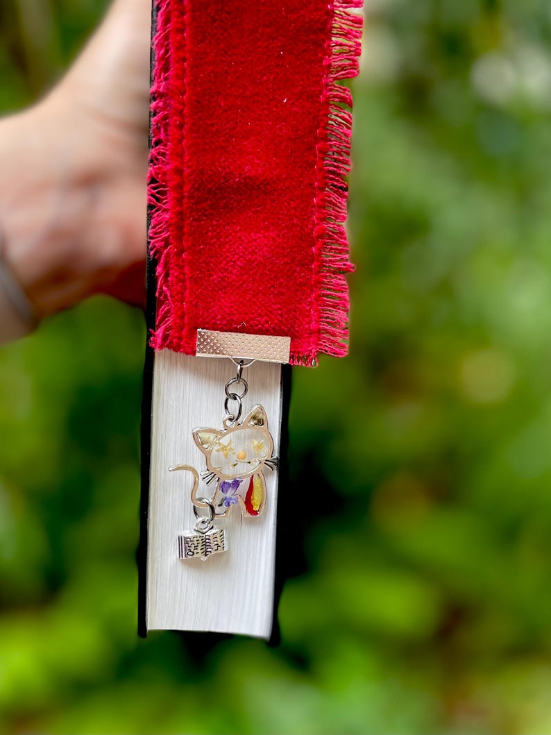 May include: A red velvet bookmark with a frayed edge and a silver charm of a cat holding a book. The cat is decorated with pressed flowers.