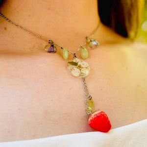 May include: A silver chain necklace with a delicate pendant featuring a white flower encased in resin. The pendant is attached to a small red strawberry charm.