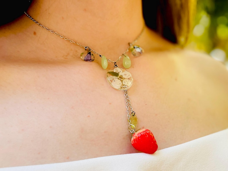 May include: A silver chain necklace with a delicate pendant featuring a white flower encased in resin. The pendant is attached to a small red strawberry charm.