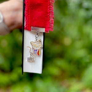 May include: A red velvet bookmark with a frayed edge and a silver charm of a cat holding a book. The cat is made of clear resin with dried flowers inside.
