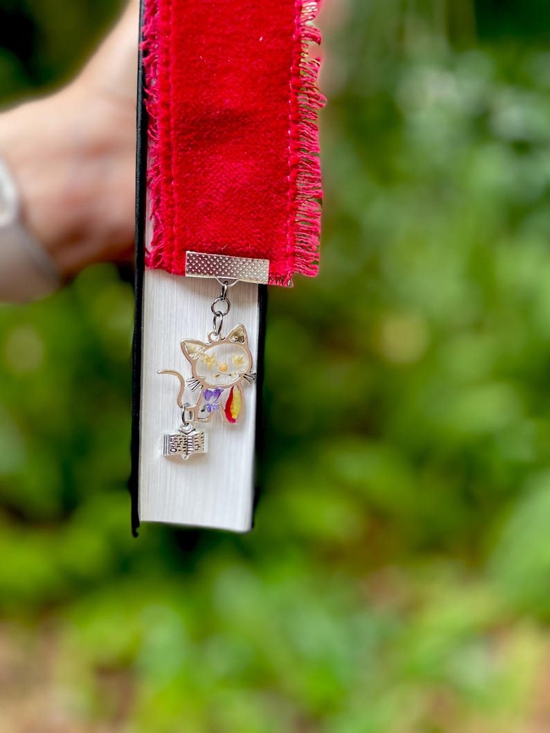 May include: A red velvet bookmark with a frayed edge and a silver charm of a cat holding a book. The cat is made of clear resin with dried flowers inside.