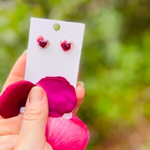 May include: A pair of heart-shaped earrings with pressed rose petals and gold flakes inside. The earrings are on a white card and are being held by a hand.