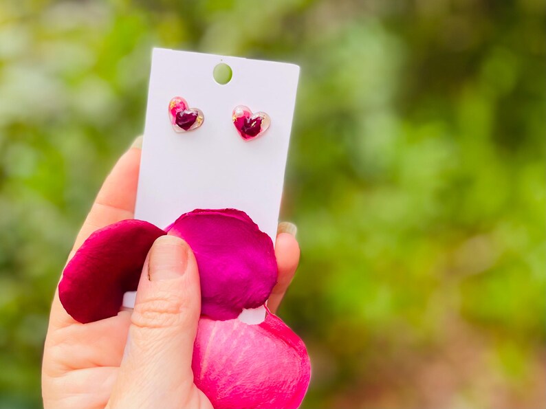May include: A pair of heart-shaped earrings with pressed rose petals and gold flakes inside. The earrings are on a white card and are being held by a hand.