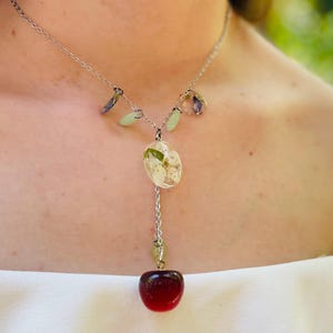 May include: A silver necklace with a red cherry charm and a pendant featuring white flowers pressed in clear resin.