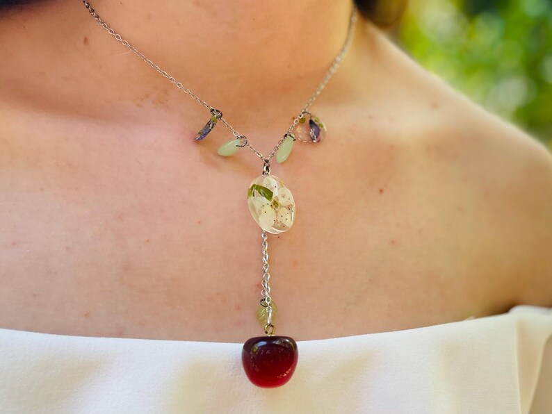 May include: A silver necklace with a red cherry charm and a pendant featuring white flowers pressed in clear resin.