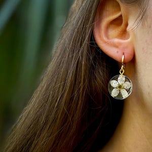 Puede incluir: Un par de pendientes de aro dorados con una flor blanca prensada dentro de un c&iacute;rculo de resina transparente.