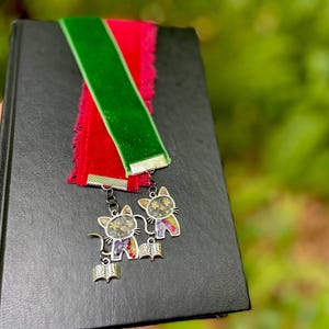 May include: A black book with a red and green velvet bookmark with two silver cat charms attached. The charms have a floral design and a book shape.