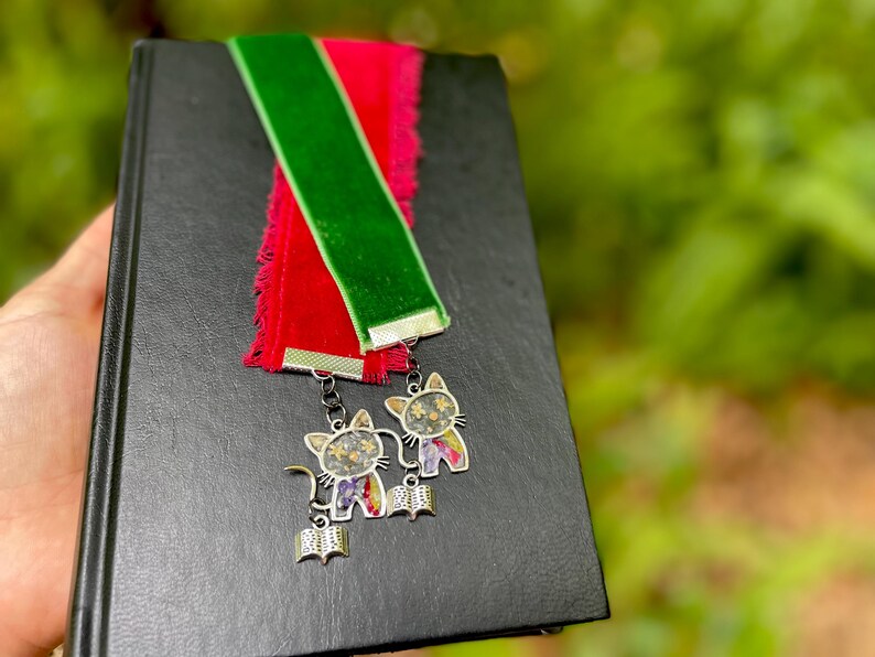 May include: A black book with a red and green velvet bookmark with two silver cat charms attached. The charms have a floral design and a book shape.