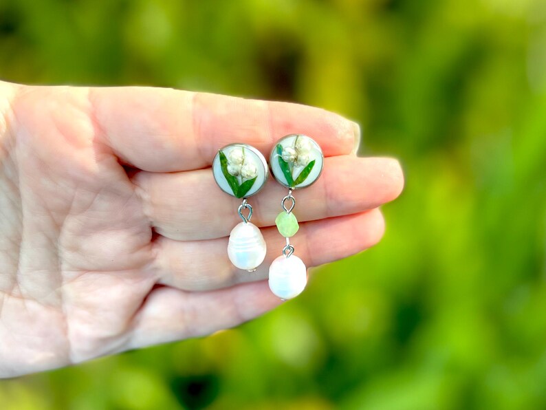 May include: Silver-toned dangle earrings featuring round, clear resin pendants with lily of the valley flowers. Each earring has a white pearl and green bead drop. The earrings are held in a hand against a blurred green background.