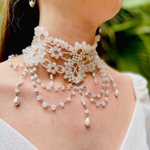 May include: A white beaded choker necklace with a delicate floral design and dangling pearl accents.