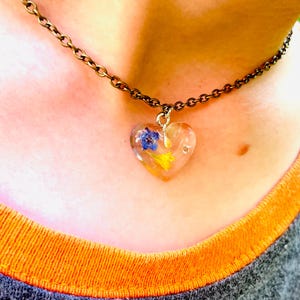 Puede incluir: Un delicado collar de cadena dorada con un colgante en forma de coraz&oacute;n lleno de flores prensadas. Las flores son azules, amarillas y blancas.
