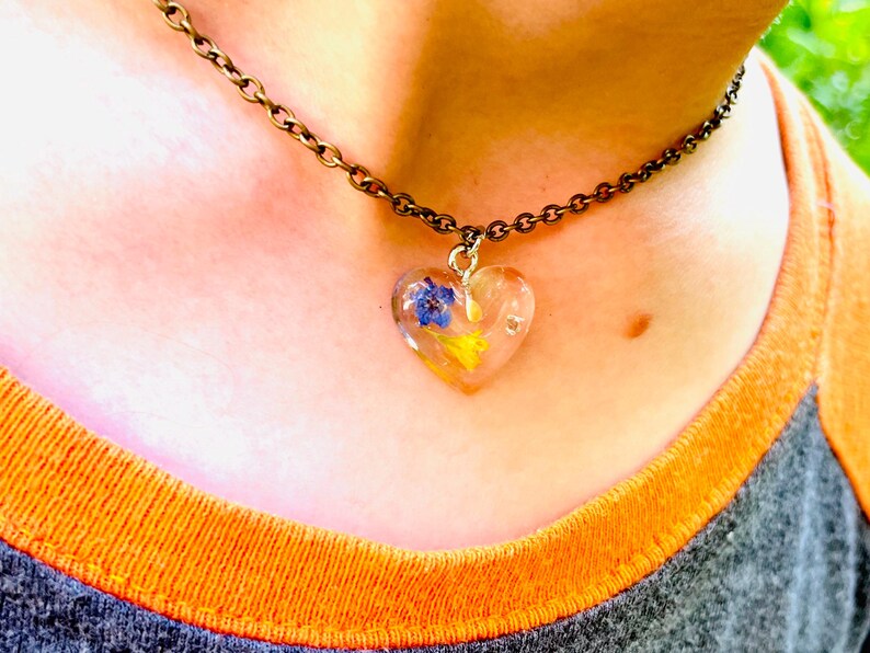 Puede incluir: Un delicado collar de cadena dorada con un colgante en forma de coraz&oacute;n lleno de flores prensadas. Las flores son azules, amarillas y blancas.