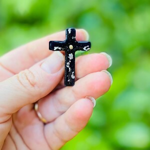 May include: A black cross with silver glitter accents and a gold dot in the center. The cross is being held in a hand.