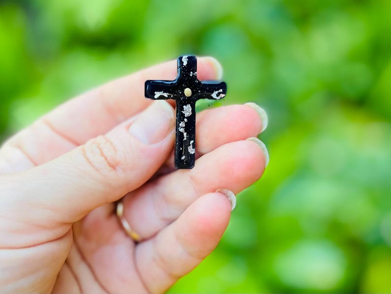 May include: A black cross with silver glitter accents and a gold dot in the center. The cross is being held in a hand.
