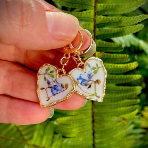 May include: A pair of gold heart-shaped earrings with pressed flowers inside. The flowers are blue and white and are set in clear resin. The earrings are on a white background.