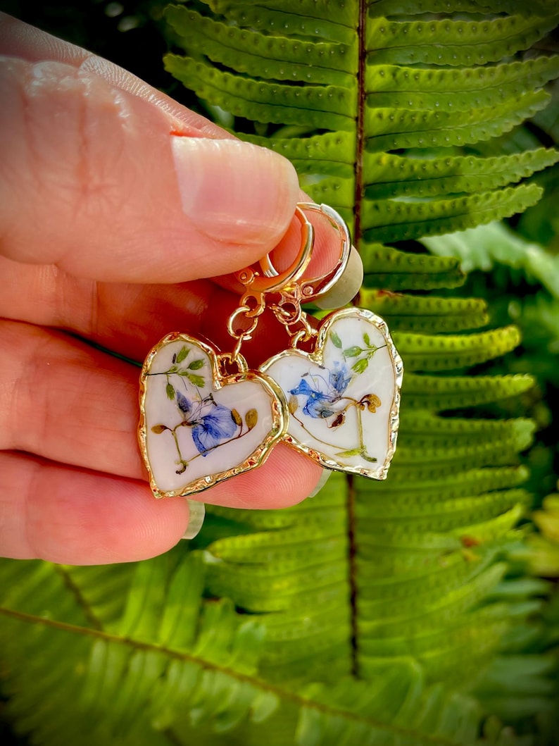 May include: A pair of gold heart-shaped earrings with pressed flowers inside. The flowers are blue and white and are set in clear resin. The earrings are on a white background.