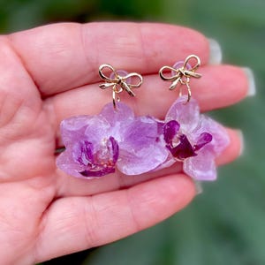 Pode incluir: Um par de brincos pendentes com flores de orqu&iacute;dea preservadas em tom lil&aacute;s claro e centros em magenta profundo. Cada brinco est&aacute; preso a um pequeno pino de metal dourado polido em forma de la&ccedil;o, criando um design floral delicado e brilhante.