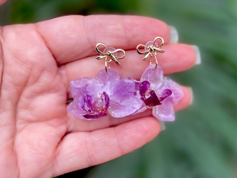 Pode incluir: Um par de brincos pendentes com flores de orqu&iacute;dea preservadas em tom lil&aacute;s claro e centros em magenta profundo. Cada brinco est&aacute; preso a um pequeno pino de metal dourado polido em forma de la&ccedil;o, criando um design floral delicado e brilhante.