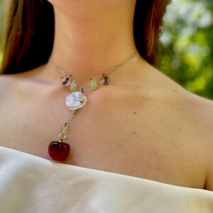 May include: A silver necklace with a red heart charm, a round white charm, and green and purple beads.
