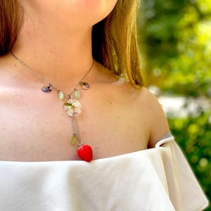 May include: A silver necklace with a delicate chain and a pendant featuring a small, red strawberry charm. The pendant also includes a round, clear resin charm with dried flowers and green and purple accents.