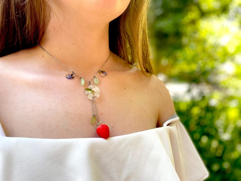 May include: A silver necklace with a delicate chain and a pendant featuring a small, red strawberry charm. The pendant also includes a round, clear resin charm with dried flowers and green and purple accents.