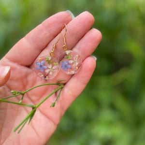 Puede incluir: Un par de pendientes de oro con resina transparente y flores prensadas. Las flores son azules y blancas.