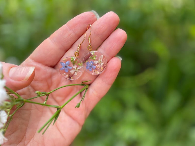 Puede incluir: Un par de pendientes de oro con resina transparente y flores prensadas. Las flores son azules y blancas.