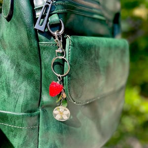 Peut inclure: Un sac &agrave; dos en cuir vert avec une fermeture &eacute;clair argent&eacute;e et un porte-cl&eacute;s contenant un charm en forme de fraise et un charm en r&eacute;sine transparente avec une fleur blanche. Le sac &agrave; dos a une finition textur&eacute;e.